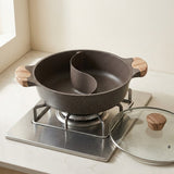 Dark speckled divided hot pot with wood-look handles sits on a gas stovetop beside a glass lid on a white countertop.