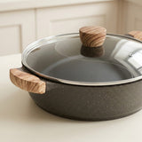 A dark gray speckled non-stick pan with a clear glass lid and light brown wooden handles, shown on a light beige countertop with a soft-focus kitchen background.