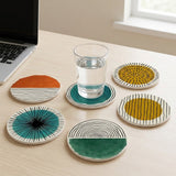 Five round ceramic coasters with abstract hand-painted patterns in orange, teal, yellow, and green, arranged on a light wood desk next to a glass of water and a laptop.