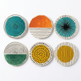 Six round coasters with mid-century modern abstract designs, arranged in a 2x3 grid on a white background. The designs feature teal, orange, green, yellow, black, and white patterns including concentric circles, sunsets, and radiating lines.