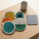 Five round coasters with colourful abstract patterns and one grey square coaster sit beside a beige cup on a light wooden table.