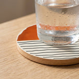 A water glass covered in condensation rests on an orange and cream coaster with black wavy lines on a wooden table.