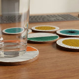 A tall clear glass rests on an orange striped coaster, with five colorful coasters and a keyboard behind it on a wooden desk.
