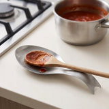 A stainless steel kitchen spoon rest holding a wooden spoon with tomato sauce, set on a light beige countertop with a pot of sauce and a gas stove in the background.