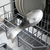 A stainless steel kitchen spoon with a curved, sculptural handle rests in the open lower basket of a dishwasher, with stacked plates visible in the upper rack.