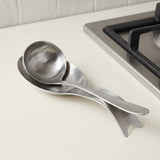 Two stainless steel kitchen utensils, one spoon and one spatula, resting on a white countertop next to a gas stovetop.
