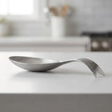 A modern stainless steel spoon-shaped dish or tray with a sculptural, wavy handle, resting on a white kitchen countertop with a blurred kitchen background.