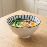 White ceramic bowl with blue interior stripes holds noodles, greens, mushrooms, and half an egg on a wooden table.
