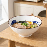 White ceramic bowl with blue cat decoration holds noodles, sliced meat, egg, seaweed, and vegetables on a wooden kitchen table.