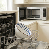 White ceramic bowl with blue interior stripes rests in an open dishwasher rack, with an open microwave on the counter behind.