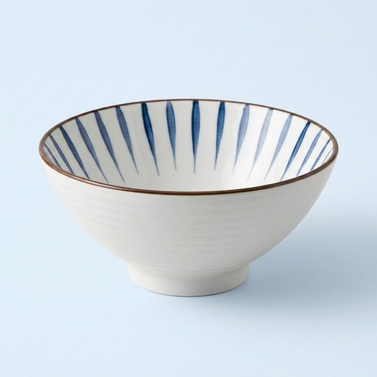 White ceramic bowl with tapered blue interior stripes and a thin brown rim sits against a pale blue background.