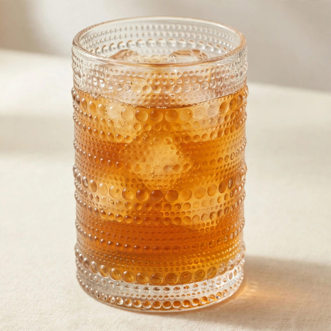 A clear textured glass tumbler with horizontal rows of raised dots, filled with an amber-colored iced drink and ice cubes, photographed on a light beige surface with a soft neutral background.