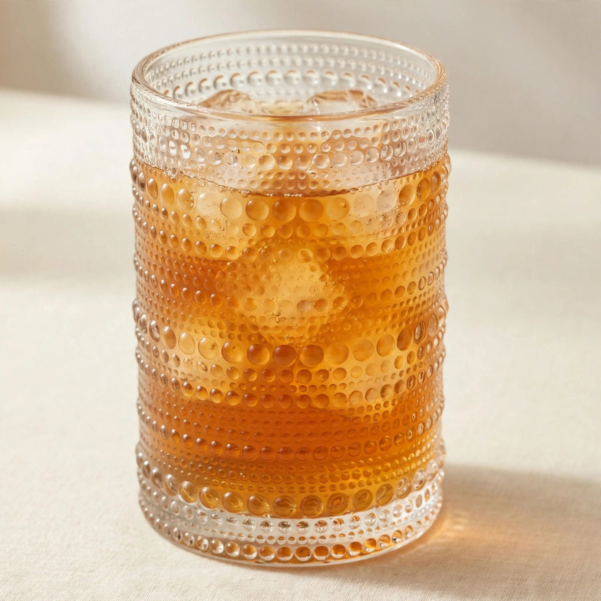A clear textured glass tumbler with horizontal rows of raised dots, filled with an amber-colored iced drink and ice cubes, photographed on a light beige surface with a soft neutral background.