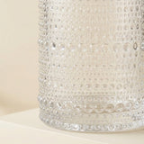 A close-up of a clear glass container with a textured, bubbly bubble pattern on a warm cream background.