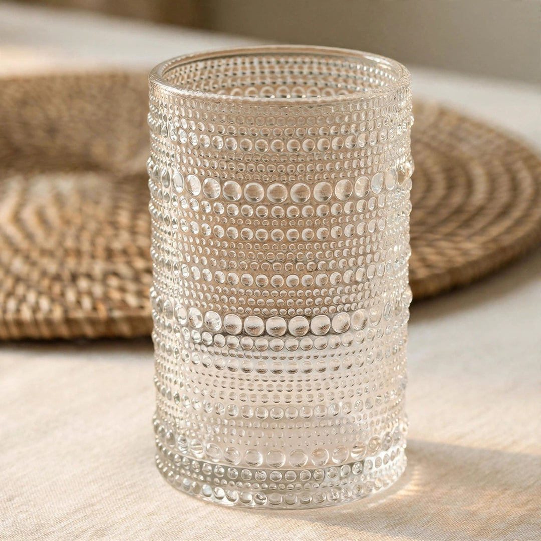 A clear textured glass tumbler with horizontal rows of raised dots, standing on a light beige surface with a woven placemat blurred in the background.