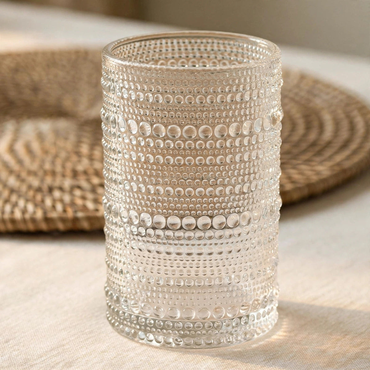 A clear textured glass tumbler with horizontal rows of raised dots, standing on a light beige surface with a woven placemat blurred in the background.