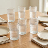 Six clear hobnail drinking glasses stand on a wooden dining table with cream plates and folded beige napkins.