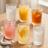 Six clear textured drinking glasses hold colourful iced drinks on a wooden table scattered with ice cubes beside a beige cloth.