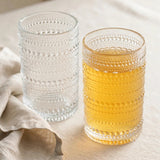 Two clear drinking glasses with raised dotted bands stand beside beige fabric, one holding a golden drink and the other clear liquid.