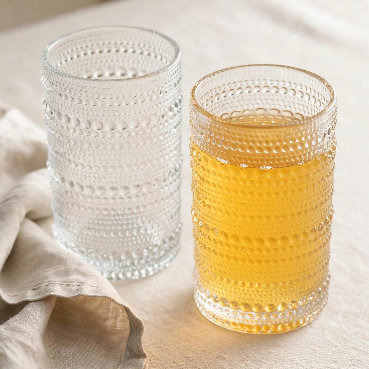 Two clear drinking glasses with raised dotted bands stand beside beige fabric, one holding a golden drink and the other clear liquid.