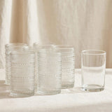 Clear textured drinking glasses stand grouped beside a single smooth glass on a sunlit pale surface against beige curtains.