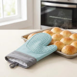 A light blue and gray silicone oven mitt resting on a baking sheet of golden-brown bread rolls on a kitchen counter, with a blurred oven and window in the background.