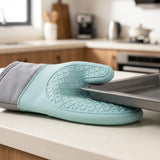 A silicone oven mitt in mint green and gray rests on a kitchen counter, holding a metal baking sheet.