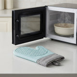 A light blue and gray silicone oven mitt with a textured grip pattern rests on a white kitchen countertop in front of an open black microwave with a bowl inside.