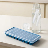 A blue silicone ice cube tray with a clear lid sits on a light beige countertop next to a clear glass bottle filled with water and ice cubes.