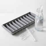 A gray ice stick tray sits beside a clear glass bottle and loose ice sticks on a white surface.