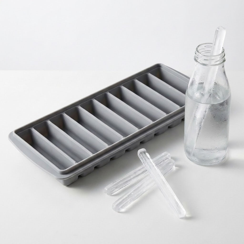 A gray ice stick tray sits beside a clear glass bottle and loose ice sticks on a white surface.