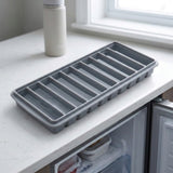 An uncovered gray ice stick tray rests on a white countertop beside a bottle and window, above an open freezer.