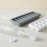 Gray and white ice trays sit beside a tipped clear plastic bottle, loose ice pieces, and a white cap on a pale surface.