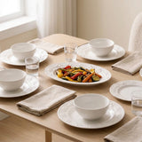 A styled dining table set with white ceramic bowls, plates, folded beige napkins, and clear drinking glasses, with a central oval platter holding colorful roasted vegetables. The scene is photographed in a bright, neutral interior with a window and curtain visible in the background.
