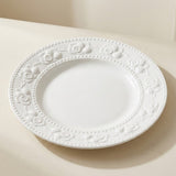 A white ceramic plate with a decorative embossed floral border, photographed on a warm beige surface with a soft shadow.