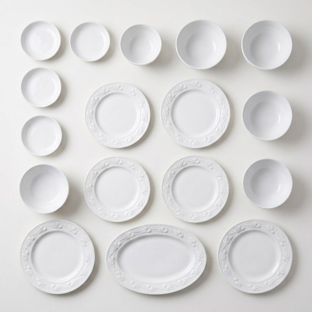 White floral embossed plates, bowls, small dishes, and an oval platter are arranged in rows on a pale background.