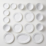 White floral embossed plates, bowls, small dishes, and an oval platter are arranged in rows on a pale background.