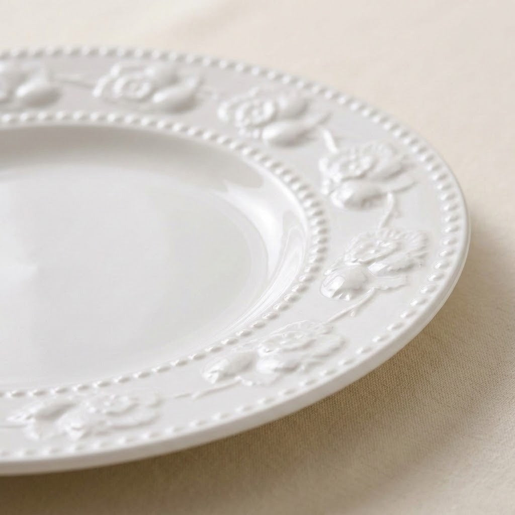 A close-up of a white ceramic plate with an embossed floral border and beaded edge, resting on a light beige fabric surface.