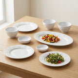 White embossed dinnerware sits on a wooden table beside a window, with vegetables on an oval platter and salad on a plate.