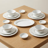 White ceramic dinnerware set arranged on a light wood table, including plates, bowls, and small cups with a subtle embossed floral pattern.