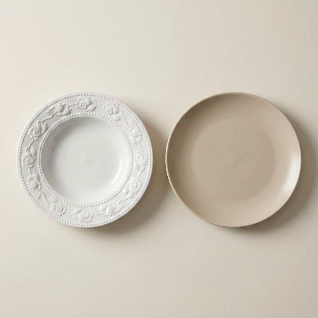 Two ceramic plates on a warm off-white background: one white plate with an embossed floral border and one matte beige plate.