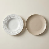Two ceramic plates on a warm off-white background: one white plate with an embossed floral border and one matte beige plate.