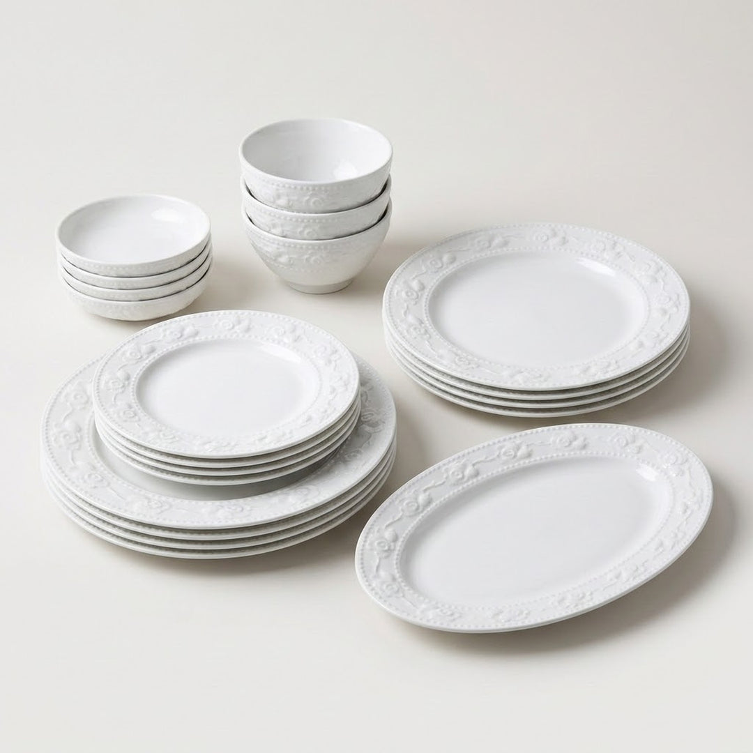 A white ceramic dinnerware set arranged on a light neutral background, including plates, bowls, and a serving platter with an embossed floral border pattern.