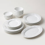 A white ceramic dinnerware set arranged on a light neutral background, including plates, bowls, and a serving platter with an embossed floral border pattern.