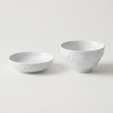 Two white ceramic bowls with embossed floral patterns, one shallow and one deeper, on a light gray background.