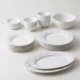 A white ceramic dinnerware set with embossed floral borders, including plates, bowls, and cups, arranged on a light beige background.