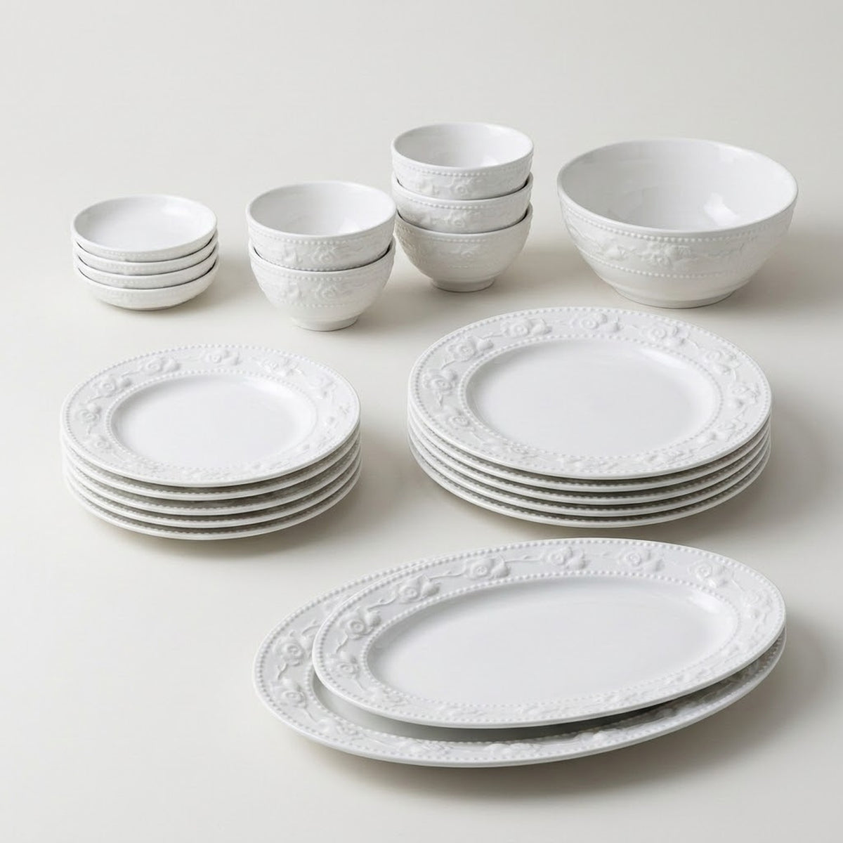 A white ceramic dinnerware set with embossed floral borders, including plates, bowls, and cups, arranged on a light beige background.