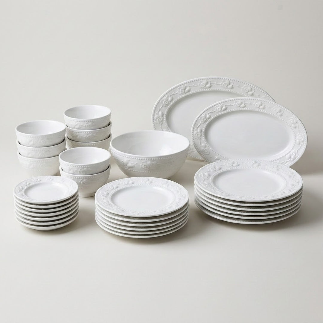 A white ceramic dinnerware set with a raised floral border pattern, including plates, bowls, and small cups, arranged neatly on a light beige background.