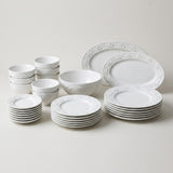 A white ceramic dinnerware set with a raised floral border pattern, including plates, bowls, and small cups, arranged neatly on a light beige background.