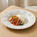A white floral embossed plate holds rice and roasted vegetables on a light wooden table.